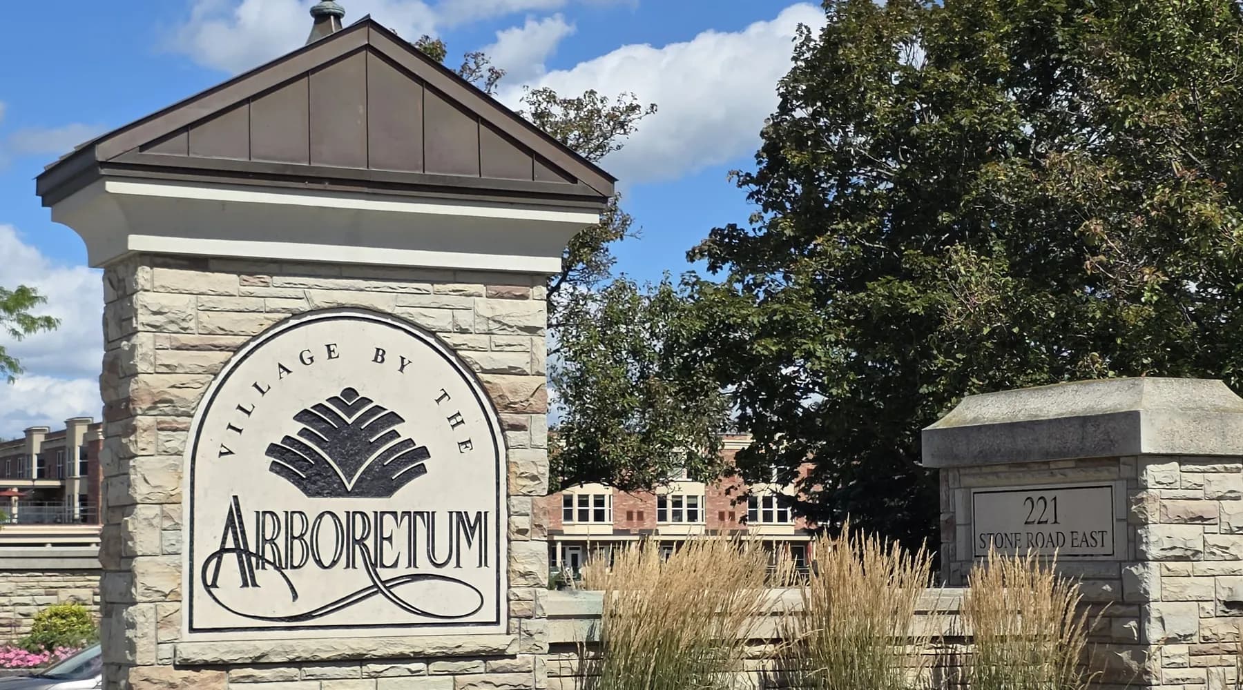 The stone entrance sign of the Village by the Arboretum at 221 Stone Road East in Guelph, with the community's buildings behind it.